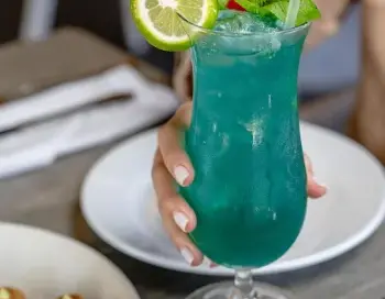 Imagen de un cóctel azul con una rodaja de lima, una ramita de menta y una baya roja en la parte superior, servido en un vaso alto y elegante. En primer plano, se aprecian dos pequeños aperitivos con una salsa amarilla y hierbas verdes esparcidas.