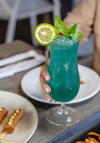 Imagen de un cóctel azul con una rodaja de lima, una ramita de menta y una baya roja en la parte superior, servido en un vaso alto y elegante. En primer plano, se aprecian dos pequeños aperitivos con una salsa amarilla y hierbas verdes esparcidas.