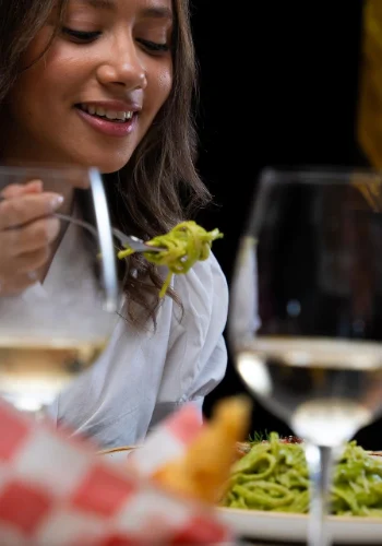 Mujer sonriente disfrutando de un plato de pasta verde en Mix Restaurant & Bar, con un tenedor en la mano. En primer plano, una copa de vino blanco y más platos en la mesa sugieren una cena elegante en este reconocido restaurante